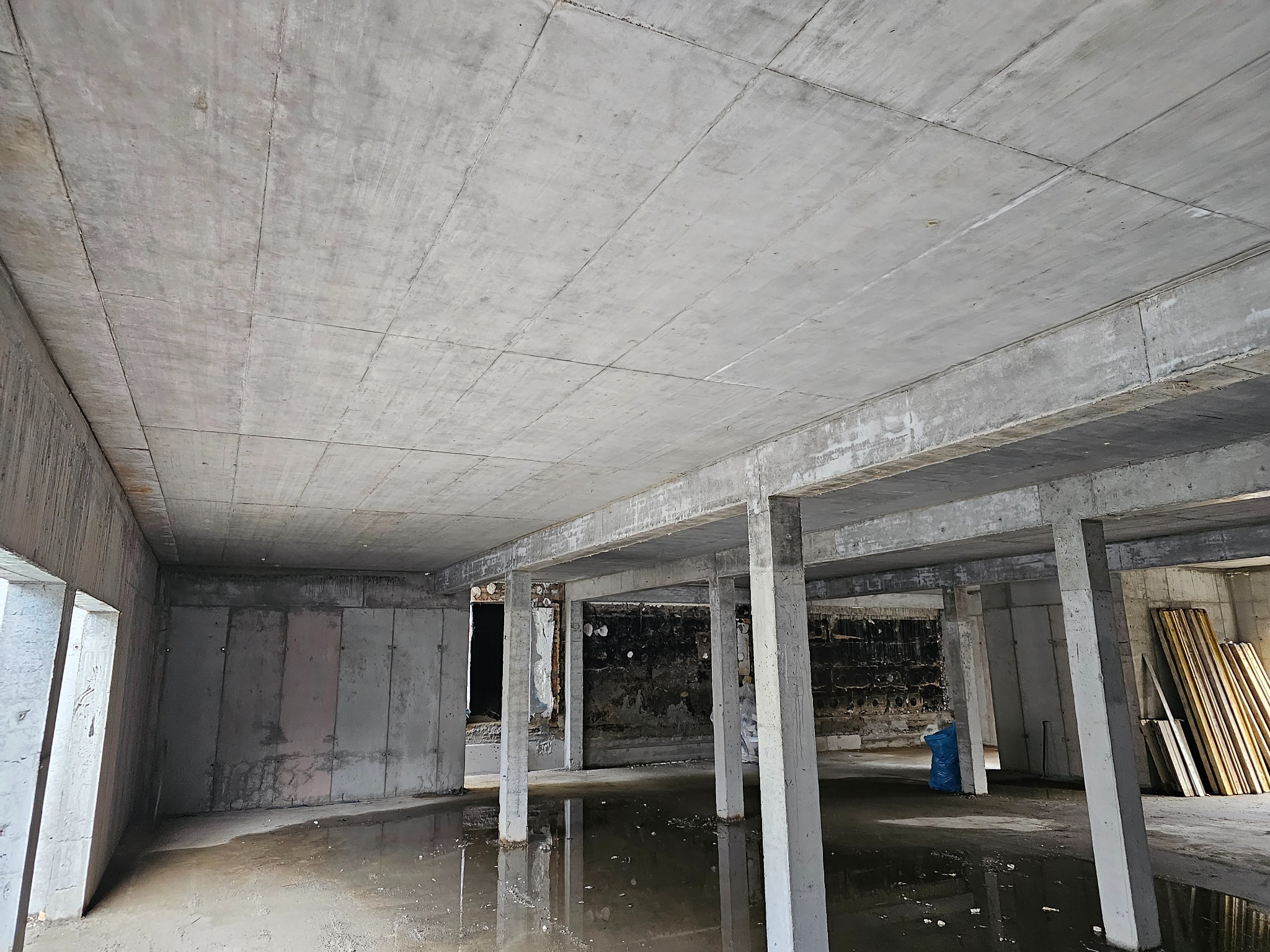 Surowy stan budynku żelbetowego z widocznymi kolumnami, stropem i ścianami. Na podłodze woda, w tle składowane deski szalunkowe. Widok z dołu na konstrukcję.