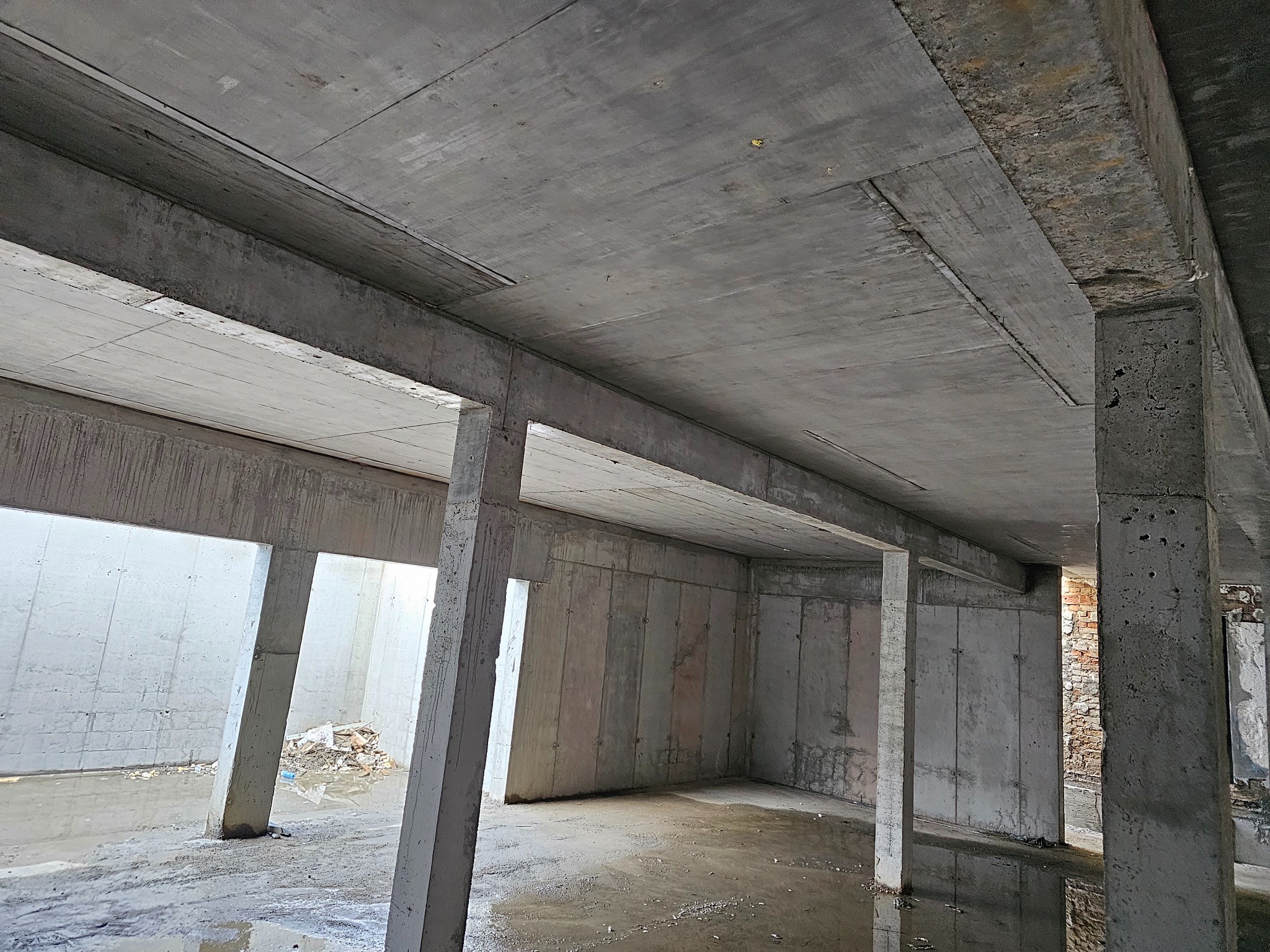 Surowy, betonowy szkielet budynku w trakcie budowy. Widoczne kolumny, ściany i strop z betonu. W tle fragment muru z cegły. Podłoże mokre, odbijające światło.