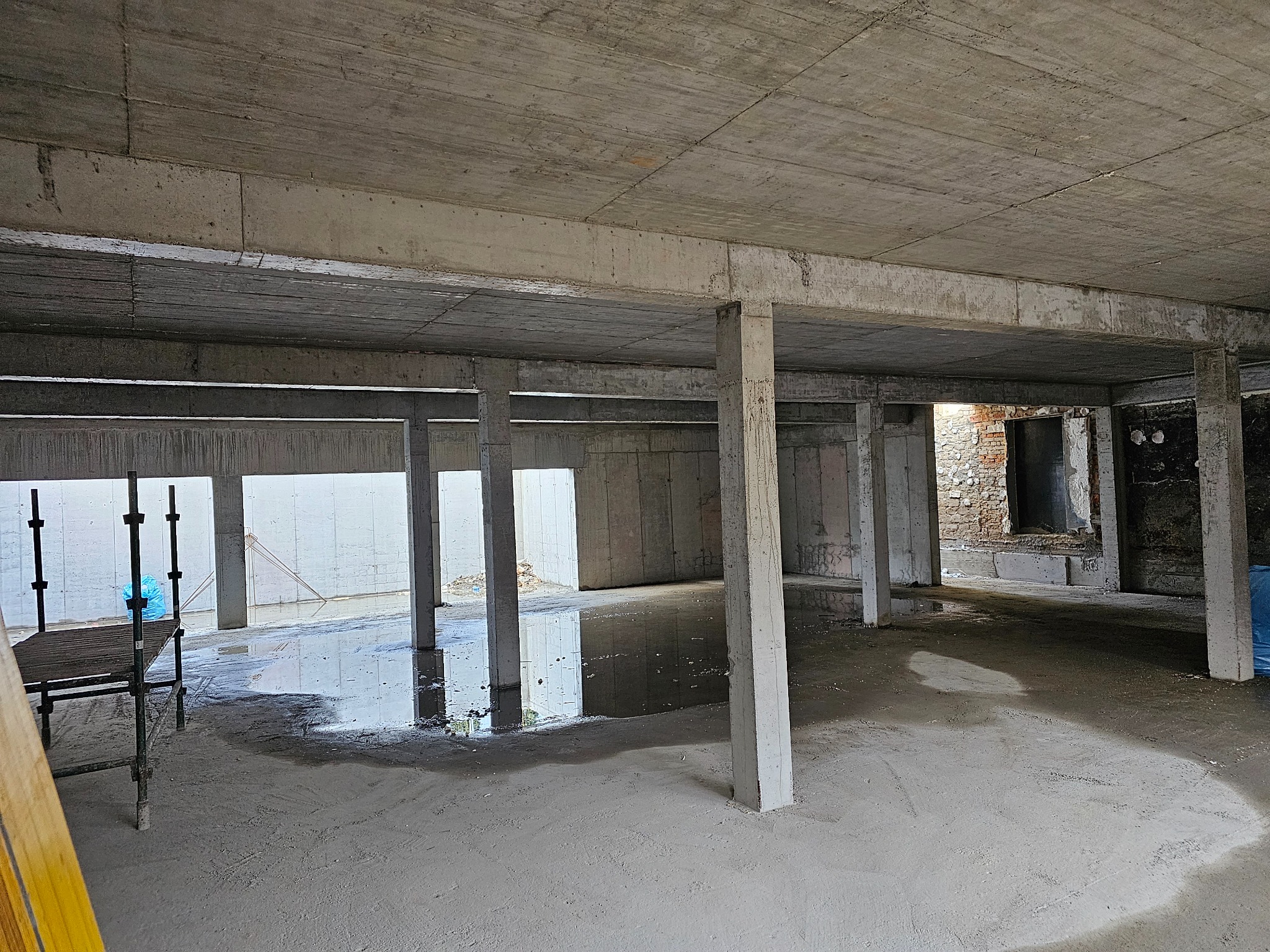 Surowy stan budynku żelbetowego: betonowy strop, kolumny i podłoga z kałużami. W tle widoczne otwory okienne i drzwiowe, a także rusztowanie.
