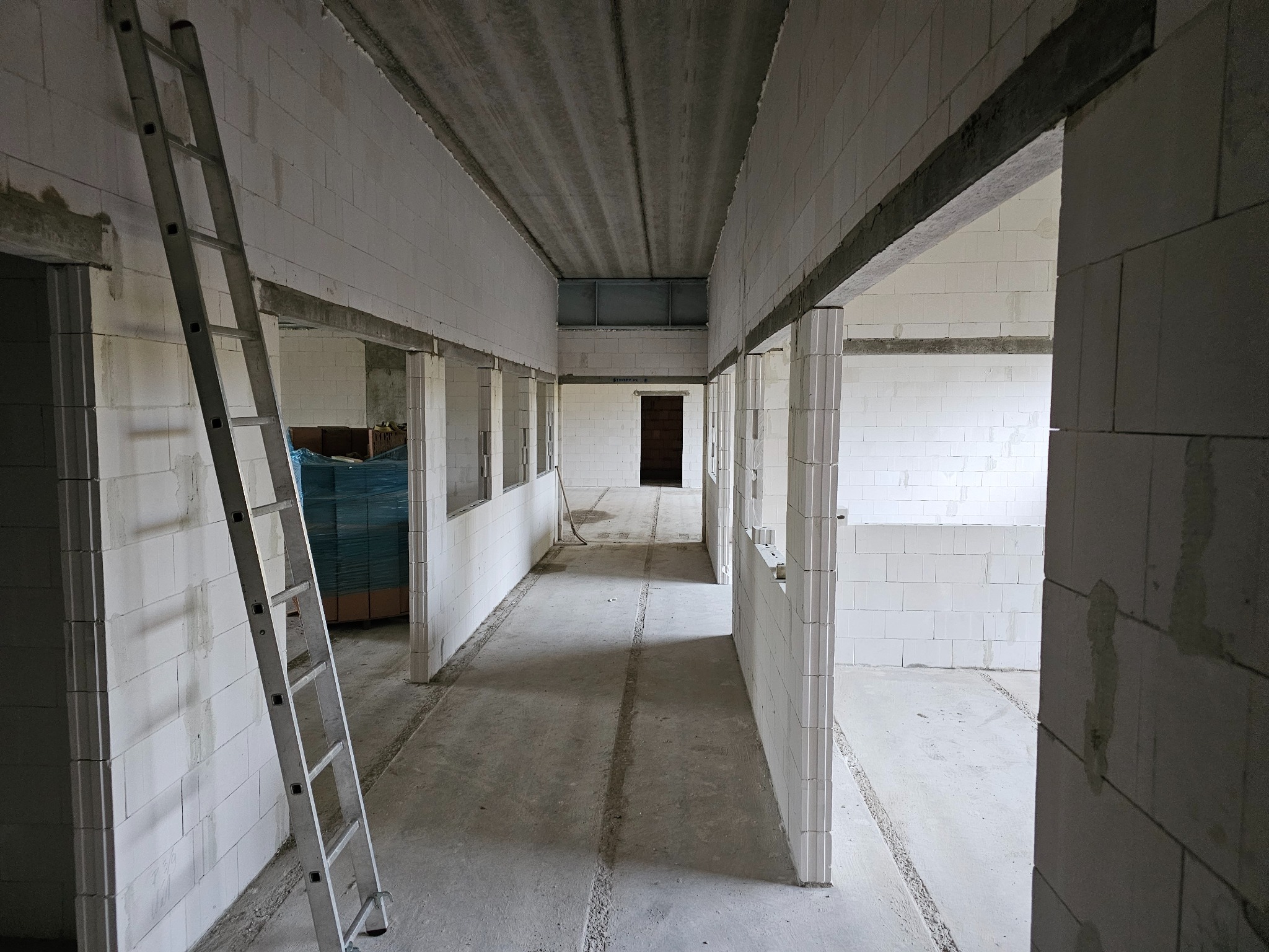 Wnętrze surowego budynku z białych bloczków, widok korytarza z drabiną opartą o ścianę. Widoczne pakiety materiałów budowlanych w tle.