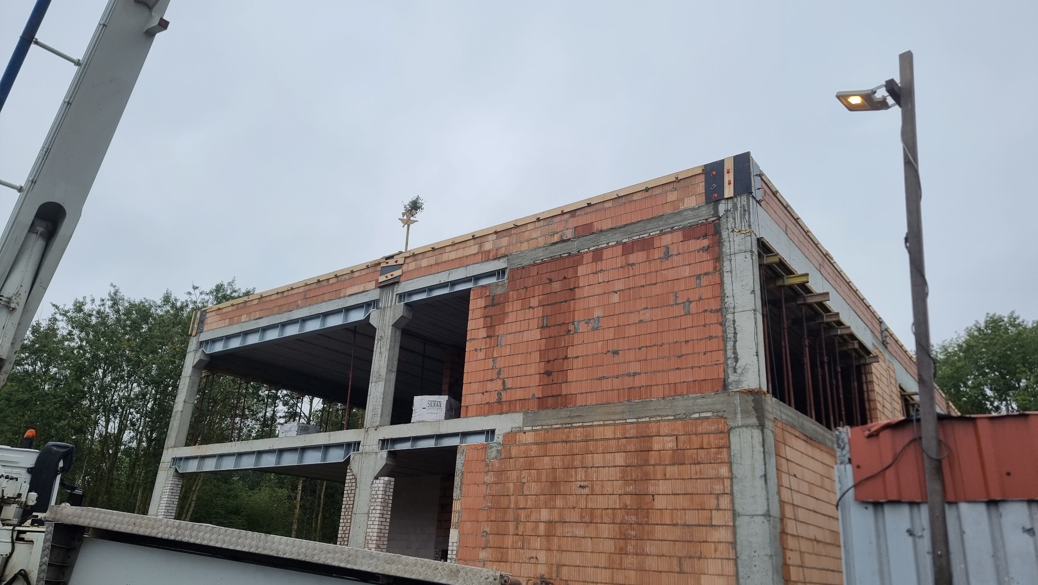 Budowa domu z czerwonej cegły, widoczne stalowe belki stropowe i betonowe słupy. Na górze budynku umieszczono małe drzewko. W tle zieleń drzew i pochmurne niebo.