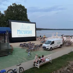 Kino plenerowe na plaży z dmuchanym ekranem wyświetlającym logo 'Mazurskie Kino Plenerowe', leżakami dla widzów, food truckiem i widokiem na jezioro w tle.