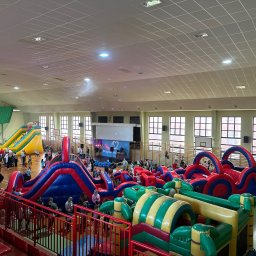 Mazurskiekinoplenerowe.pl - Wnętrze hali sportowej z rozstawionymi, kolorowymi dmuchanymi torami przeszkód i zjeżdżalniami, wypełnione uczestnikami imprezy.