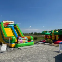 Dwa kolorowe, tematyczne place zabaw w Rucianem-Nidzie: zjeżdżalnia z motywem dinozaurów i konstrukcja w kształcie krokodyla z otwartą paszczą, ustawione na szarym bruku pod błękitnym niebem.