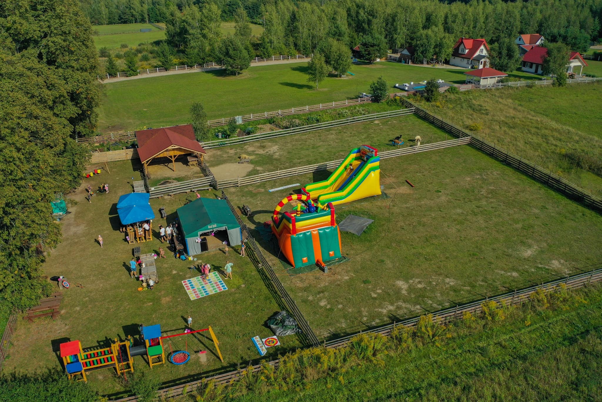 Widok z lotu ptaka na teren rekreacyjny z dmuchanym zamkiem i zjeżdżalnią, placem zabaw, altaną oraz strefą gastronomiczną otoczony zielonymi łąkami i lasem.