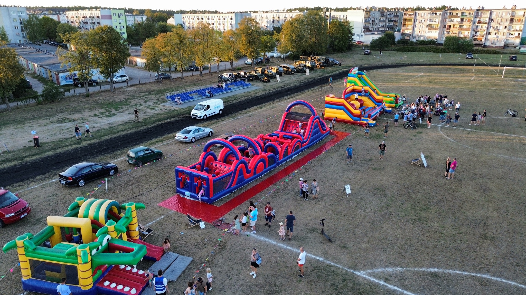 Widok z góry na rozstawione na boisku dmuchane konstrukcje: tor przeszkód, zjeżdżalnie i zamek, z uczestnikami wydarzenia w tle bloków mieszkalnych.