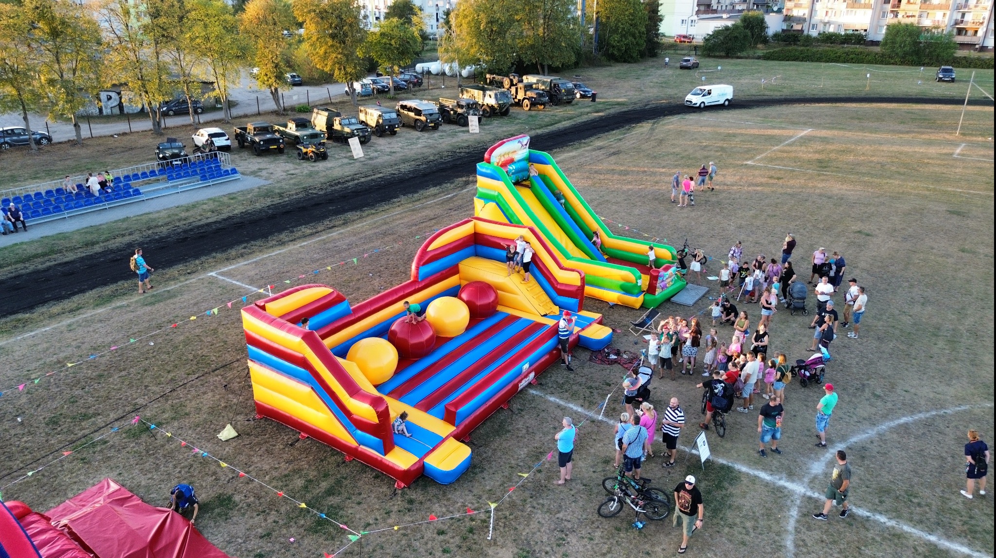 Widok z góry na rozstawione na trawiastym boisku dwa kolorowe, dmuchane zamki dla dzieci, z których jeden ma zjeżdżalnię i duże, dmuchane kule, a wokół bawią się ludzie.