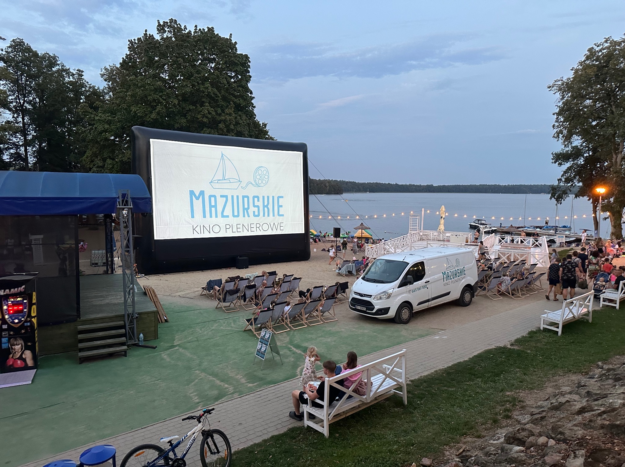 Kino plenerowe na plaży z dmuchanym ekranem wyświetlającym logo 'Mazurskie Kino Plenerowe', leżakami dla widzów, food truckiem i widokiem na jezioro w tle.