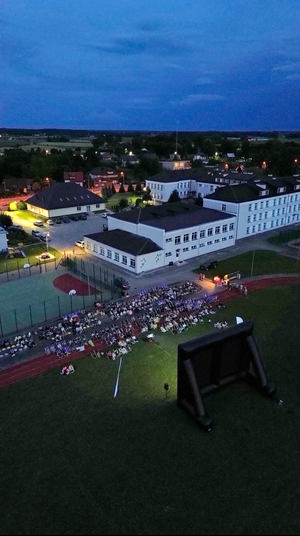Widok z lotu ptaka na plenerowe kino z dmuchanym ekranem i widownią na krzesełkach na boisku szkolnym o zmierzchu.