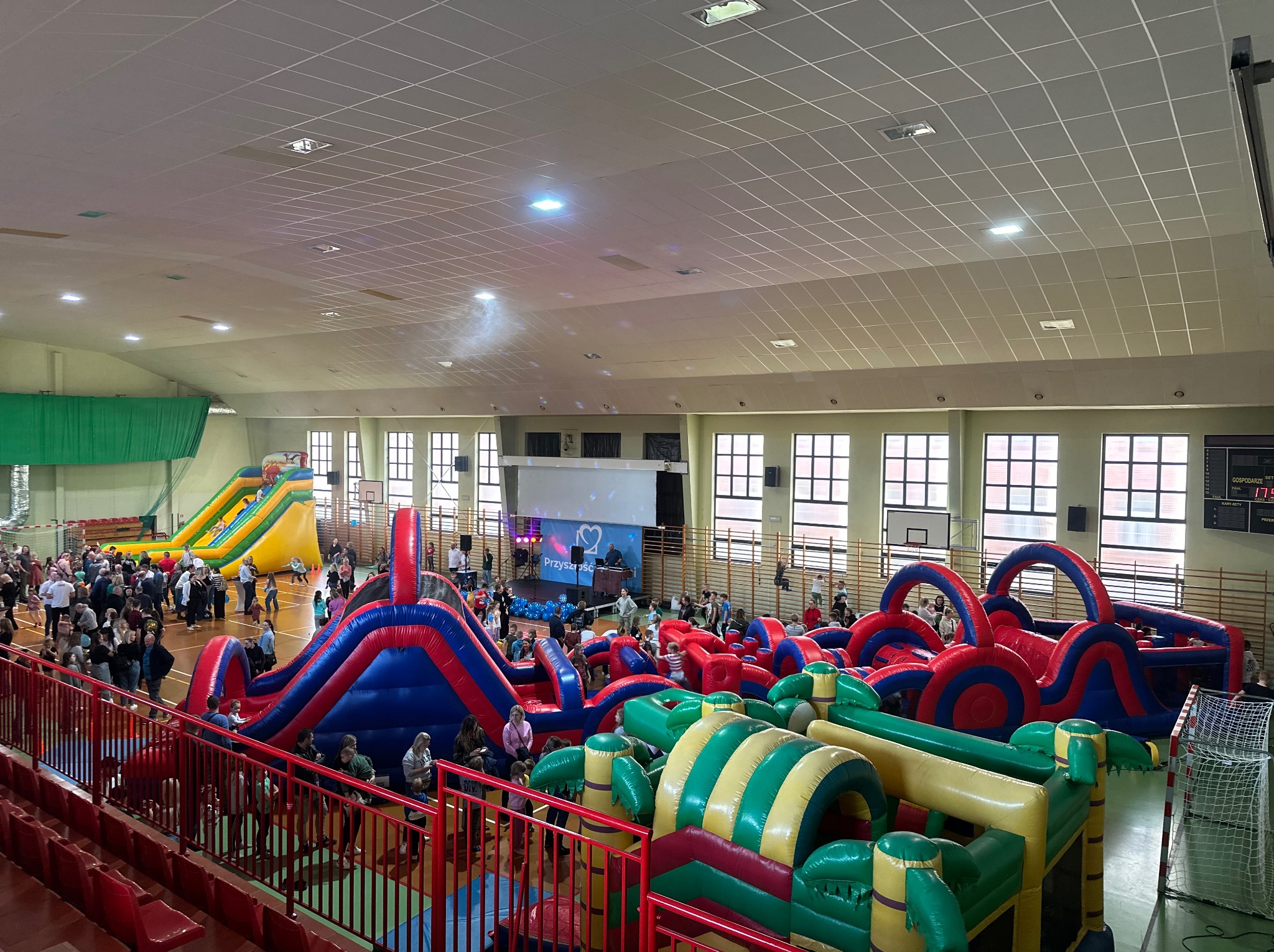 Wnętrze hali sportowej z rozstawionymi, kolorowymi dmuchanymi torami przeszkód i zjeżdżalniami, wypełnione uczestnikami imprezy.