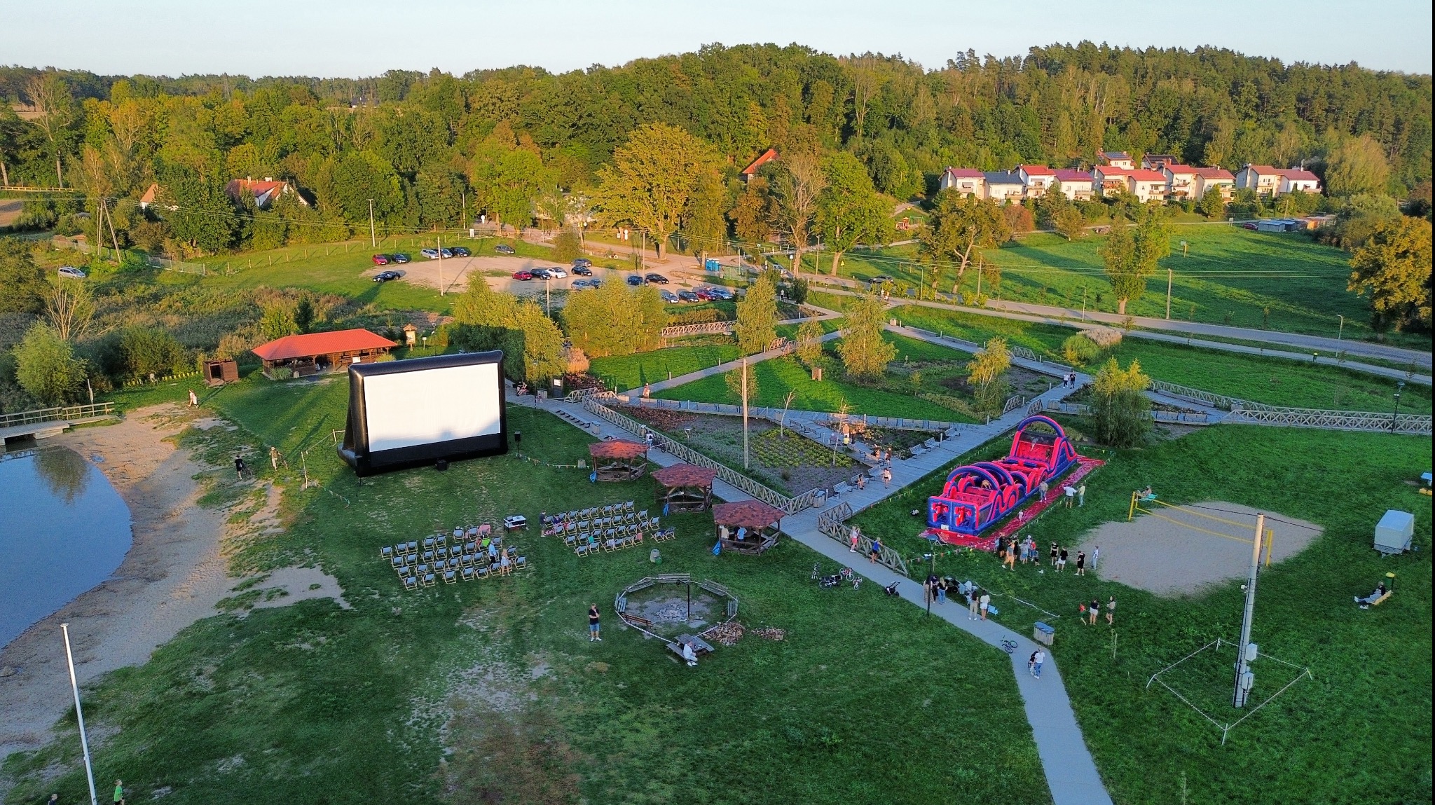 Widok z lotu ptaka na plenerowe kino z dmuchanym ekranem, rzędy krzeseł dla widzów, strefę z dmuchanym torem przeszkód, altany i ścieżki spacerowe na zielonym terenie rekreacyjnym.