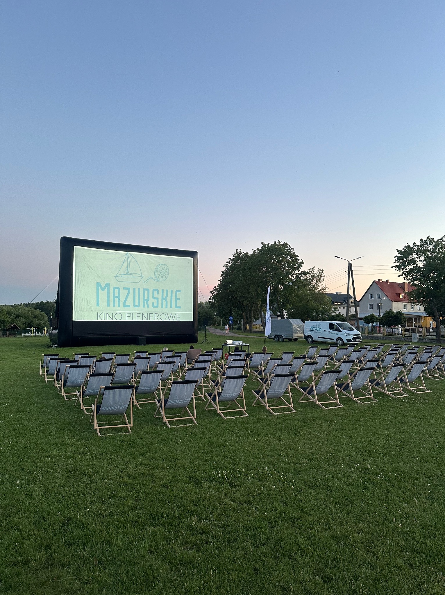 Kino plenerowe 'Mazurskie' z dmuchanym ekranem, rzędami leżaków na trawie i zaparkowanym busem w tle, gotowe na seans.