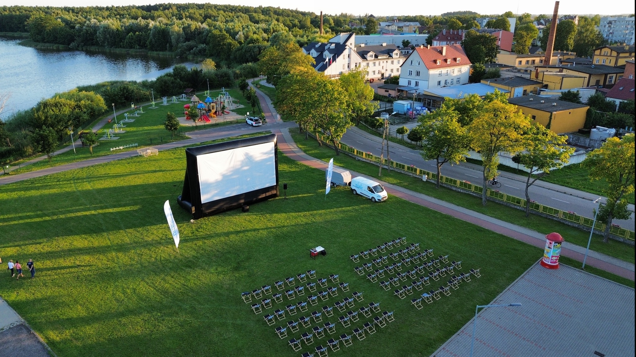 Kino plenerowe z dmuchanym ekranem, rzędami krzeseł i projektorem, z widokiem na jezioro i zabudowania miejskie w tle, ujęcie z lotu ptaka.