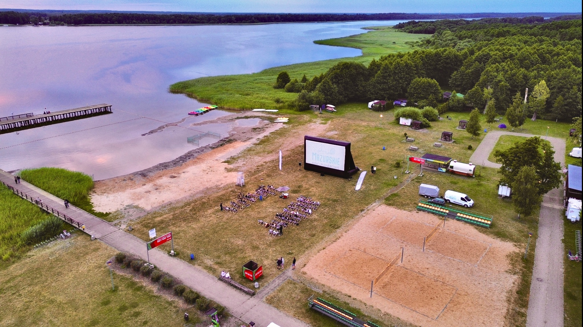 Kino plenerowe 'Mazurskie Kino Plenerowe' z dmuchanym ekranem na plaży nad jeziorem, widok z lotu ptaka, widoczny pomost, plaża, las i boisko do siatkówki.