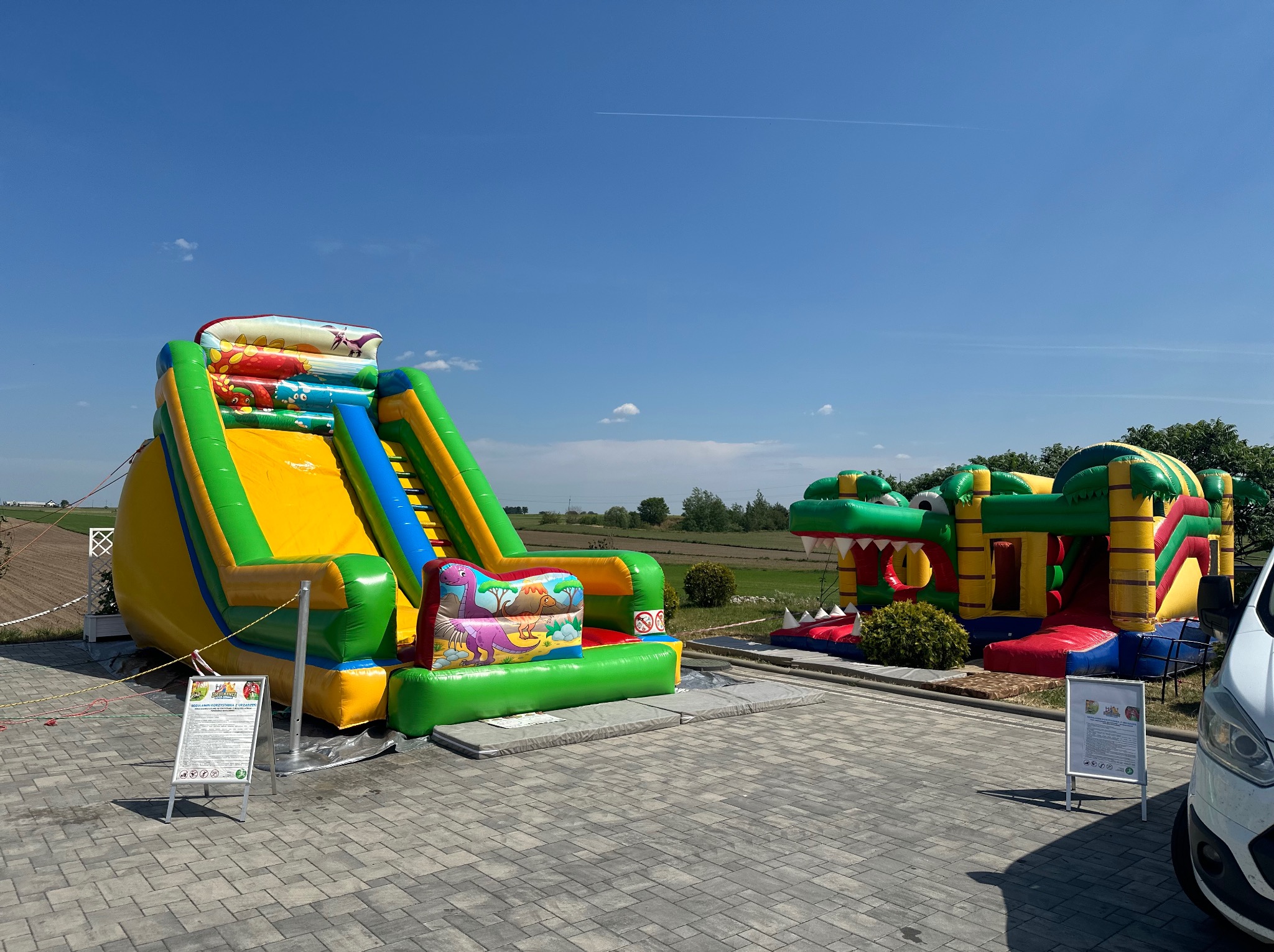Dwa kolorowe, tematyczne place zabaw w Rucianem-Nidzie: zjeżdżalnia z motywem dinozaurów i konstrukcja w kształcie krokodyla z otwartą paszczą, ustawione na szarym bruku pod błękitnym niebem.