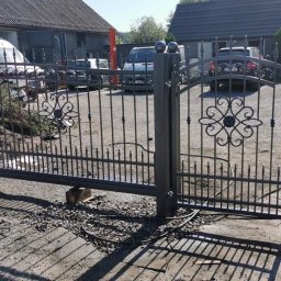 Eryk - Rozsuwana brama wjazdowa z metaloplastyki, wzór z rozetami, częściowo zamontowana, podparta drewnianymi klockami, widoczny betonowy słupek z czerwonym sznurkiem, w tle budynek gospodarczy i samochody.