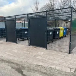 Metalowa konstrukcja z siatki i pełnymi panelami, tworząca osłony na pojemniki do segregacji śmieci w kolorach: czarnym, żółtym i niebieskim, na zewnątrz, na kostce brukowej.