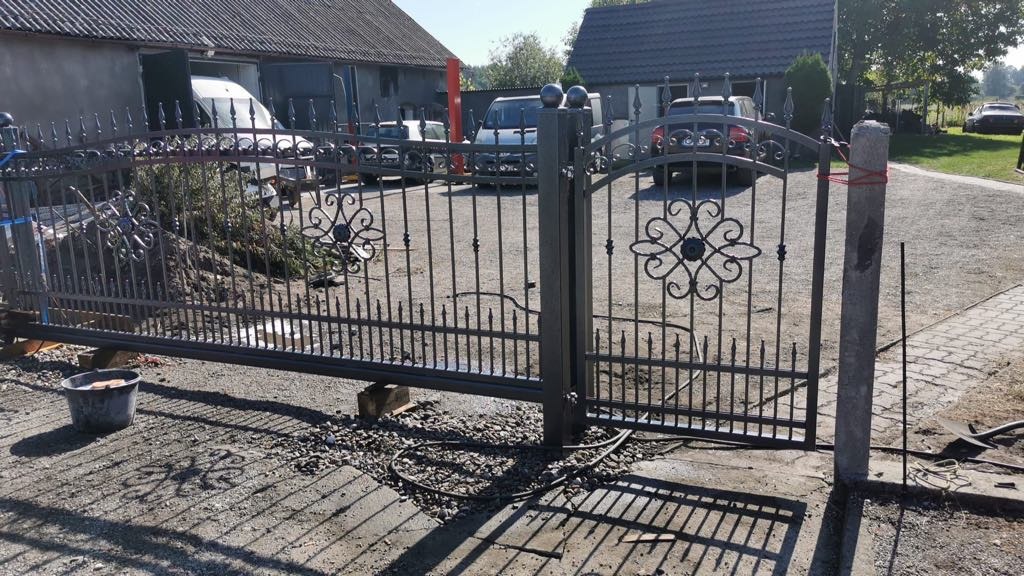 Rozsuwana brama wjazdowa z metaloplastyki, wzór z rozetami, częściowo zamontowana, podparta drewnianymi klockami, widoczny betonowy słupek z czerwonym sznurkiem, w tle budynek gospodarczy i samochody.