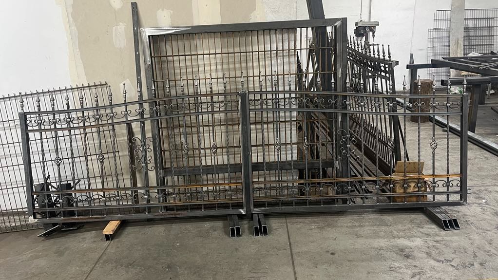 Surowe, metalowe elementy bram i ogrodzeń z dekoracyjnymi detalami, ustawione w warsztacie przed dalszą obróbką lub montażem.
