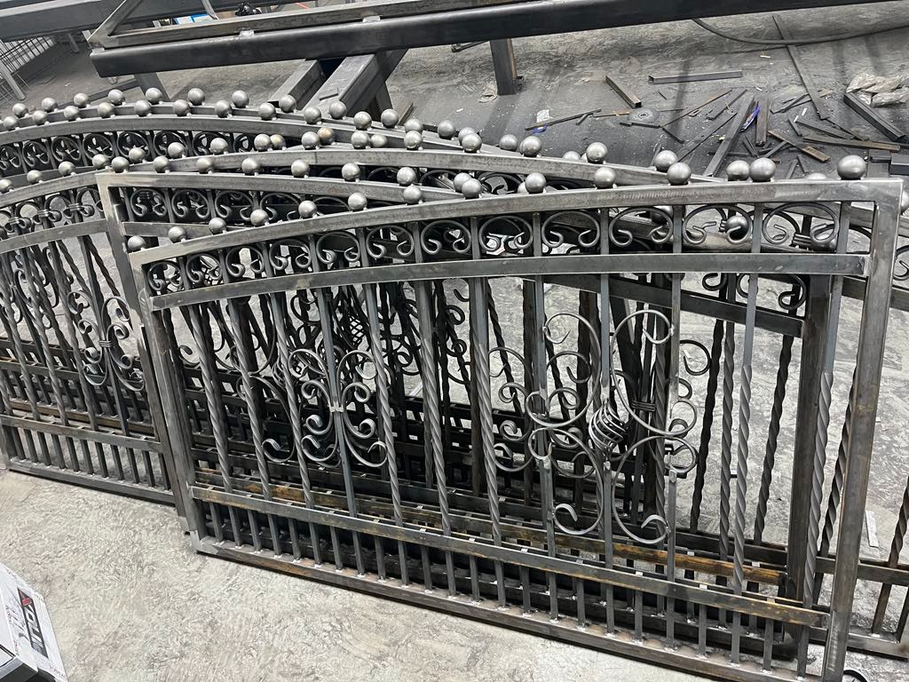 Surowe, metalowe elementy bramy z dekoracyjnymi, kręconymi wzorami i kulistymi zakończeniami, leżące na betonowej podłodze warsztatu.