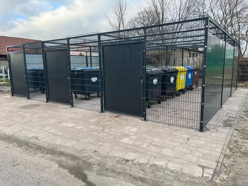 Metalowa konstrukcja z siatki i pełnymi panelami, tworząca osłony na pojemniki do segregacji śmieci w kolorach: czarnym, żółtym i niebieskim, na zewnątrz, na kostce brukowej.