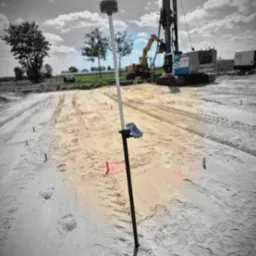 Pomiar terenu pod budowę z użyciem odbiornika GNSS RTK na tyczce. W tle maszyny budowlane i wyznaczony obszar. Praca w toku, teren przygotowywany pod inwestycję.