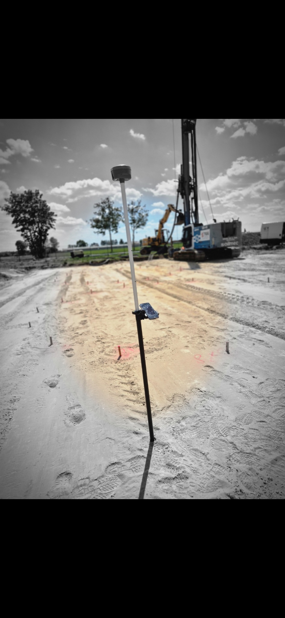 Pomiar terenu pod budowę z użyciem odbiornika GNSS RTK na tyczce. W tle maszyny budowlane i wyznaczony obszar. Praca w toku, teren przygotowywany pod inwestycję.