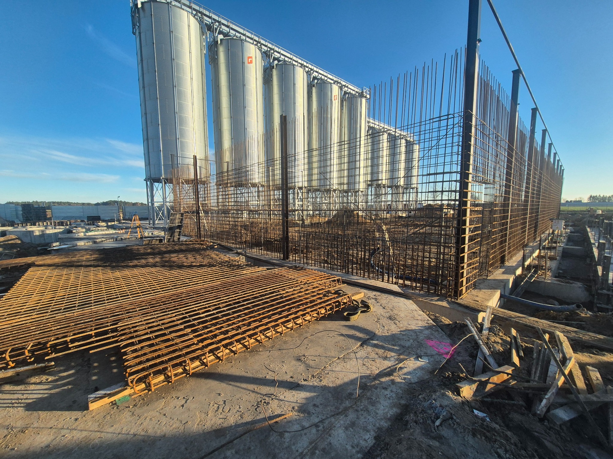 Silosy zbożowe w trakcie budowy, widoczne zbrojenia i fundamenty. Jasne niebo, wczesna faza prac ziemnych.