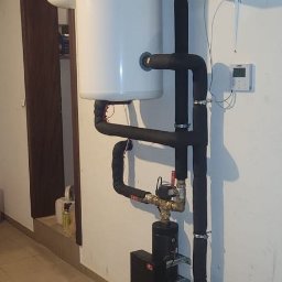 GRUPA ERMAX SP ZOO - Biały bojler z czarną izolacją rur w garażu, widoczne metalowe elementy i regulator temperatury na ścianie. Instalacja grzewcza w piwnicy.