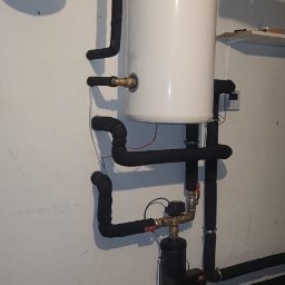 GRUPA ERMAX SP ZOO - Biały zbiornik akumulacyjny ciepła z czarnymi rurami w izolacji, połączony z pompą obiegową. Instalacja w kotłowni, widoczne elementy mocujące do ściany.