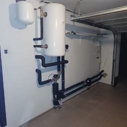 GRUPA ERMAX SP ZOO - Biały zbiornik buforowy z czarnymi rurami w garażu, element instalacji grzewczej po montażu, widok z dołu, płytki na podłodze.