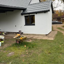 GRUPA ERMAX SP ZOO - Biała elewacja domu z ciemną stolarką okienną i dachem. W tle wózek z dyniami, trawnik i budynek gospodarczy. Jesienna sceneria.