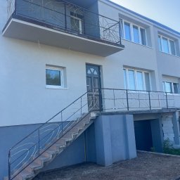 GRUPA ERMAX SP ZOO - Dwukondygnacyjny dom z balkonem i zewnętrznymi schodami z ozdobnymi, metalowymi balustradami. Elewacja w odcieniach szarości, okna z białymi ramami. Ujęcie z dołu.