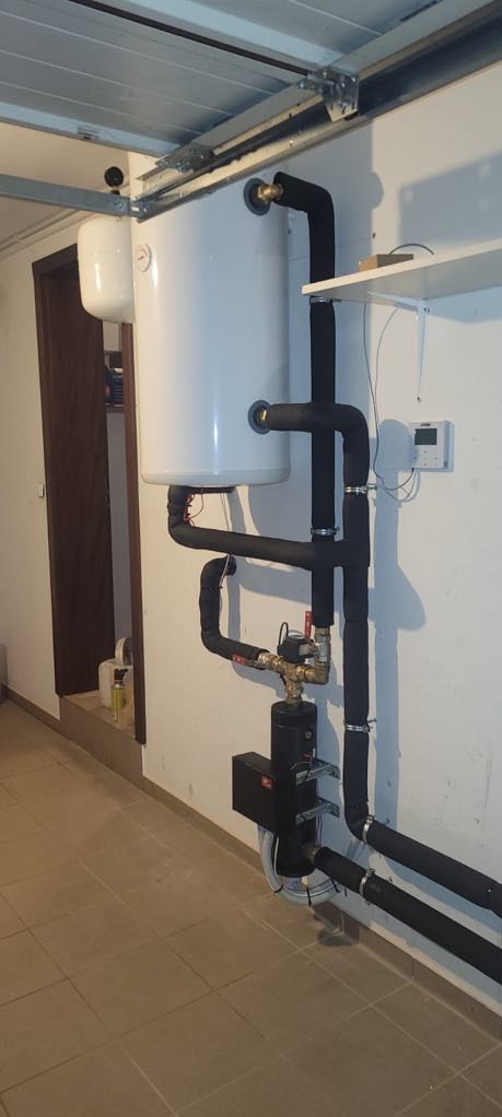 Biały bojler z czarną izolacją rur w garażu, widoczne metalowe elementy i regulator temperatury na ścianie. Instalacja grzewcza w piwnicy.