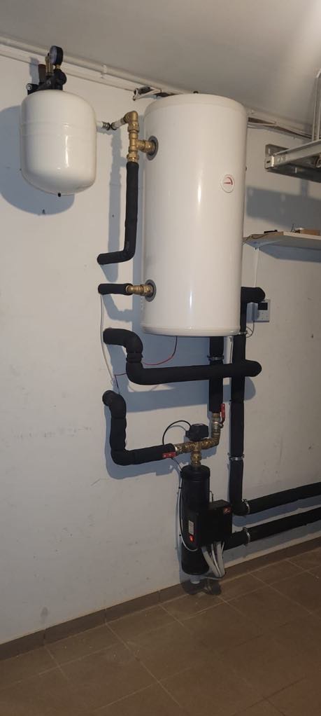 Biały zbiornik akumulacyjny ciepła z czarnymi rurami w izolacji, połączony z pompą obiegową. Instalacja w kotłowni, widoczne elementy mocujące do ściany.