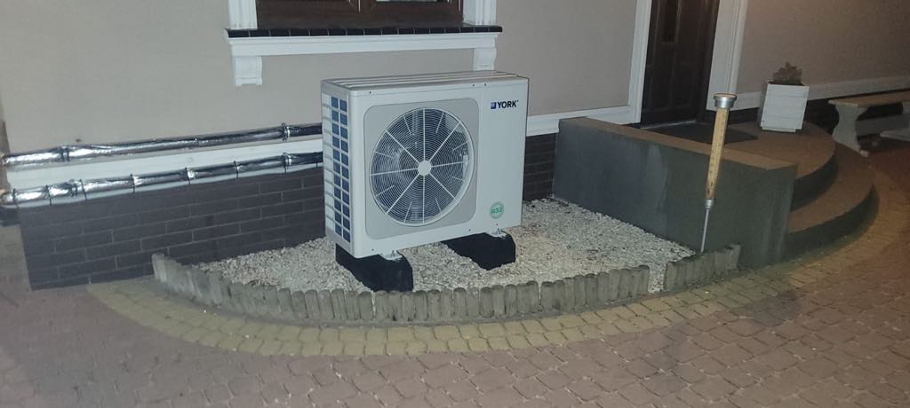 Zewnętrzna jednostka klimatyzacji YORK na tle elewacji domu, widoczne rury w izolacji. Otoczenie jednostki wyłożone drobnymi kamieniami i obrzeżem.