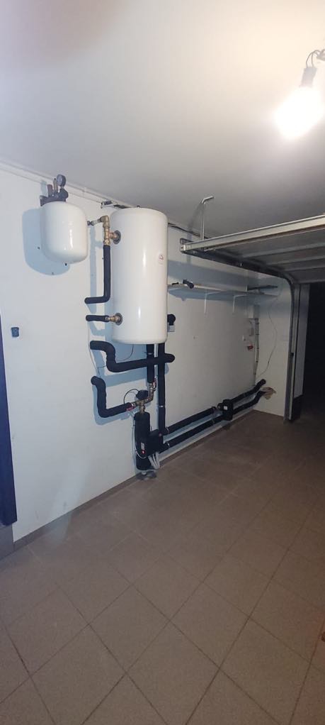 Biały zbiornik buforowy z czarnymi rurami w garażu, element instalacji grzewczej po montażu, widok z dołu, płytki na podłodze.