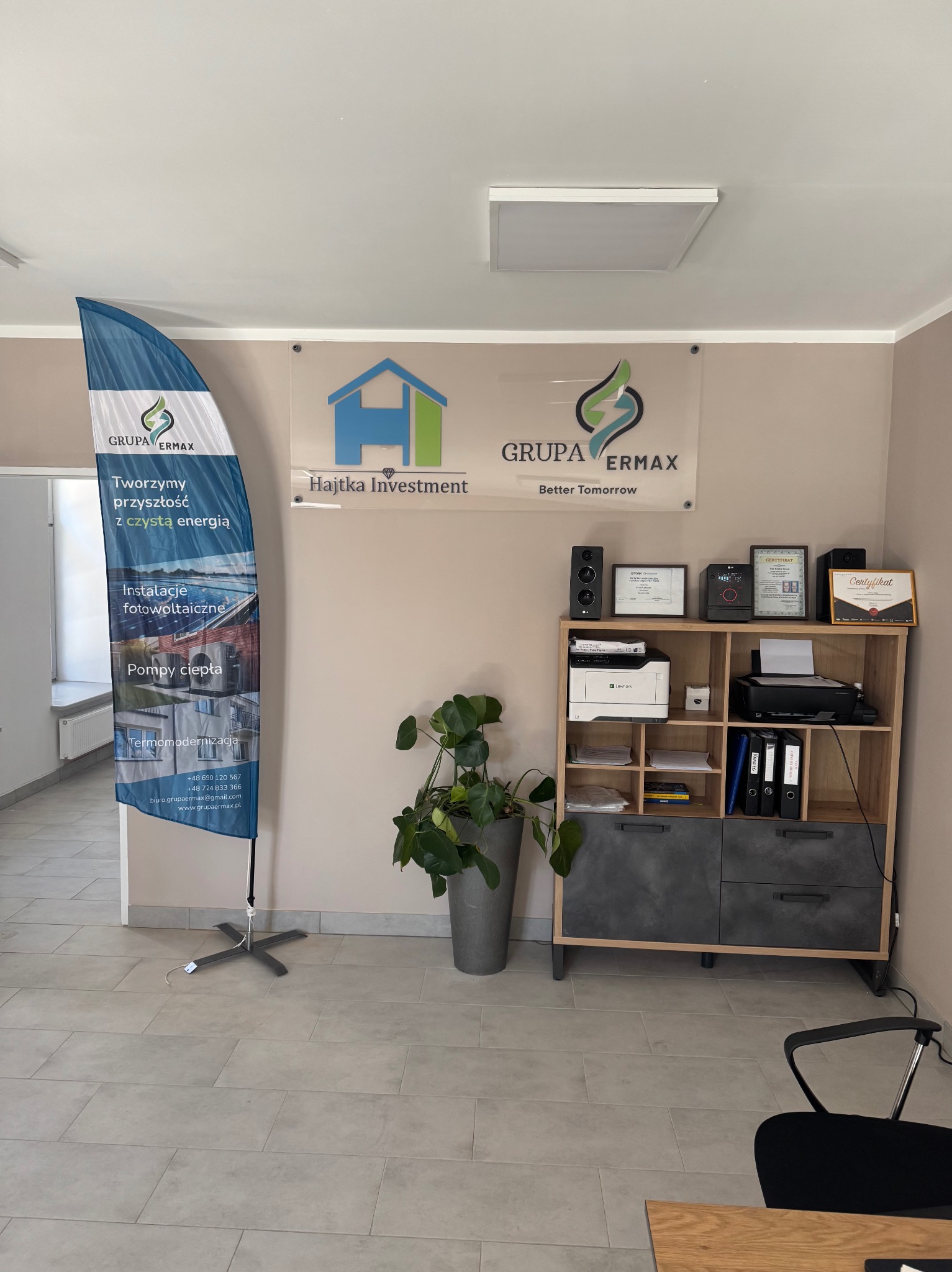Wnętrze biura firmy Grupa Ermax z flagą reklamową, certyfikatami i logo Hajtka Investment. Widoczne instalacje fotowoltaiczne, pompy ciepła i termomodernizacja.