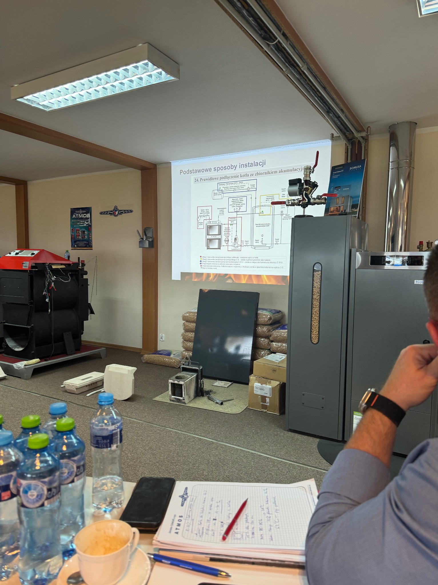 Szkolenie z instalacji kotłów: prezentacja schematu podłączenia kotła ze zbiornikiem akumulacyjnym, kotły na pellet i węgiel, notatki, butelki z wodą.