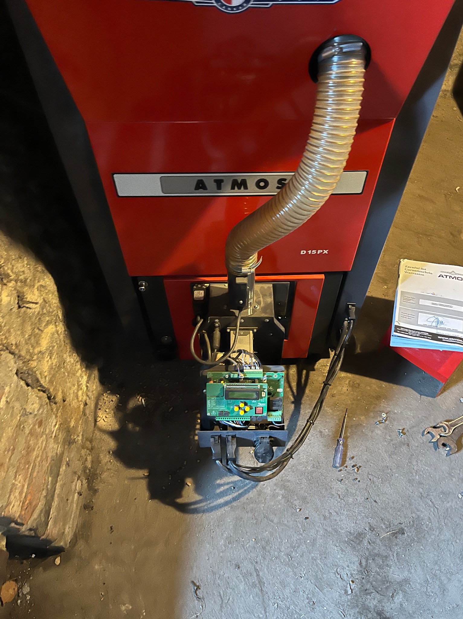 Czerwony piec Atmos D15PX z podłączonym sterownikiem i rurą, widoczne narzędzia i dokumentacja, ujęcie z góry.