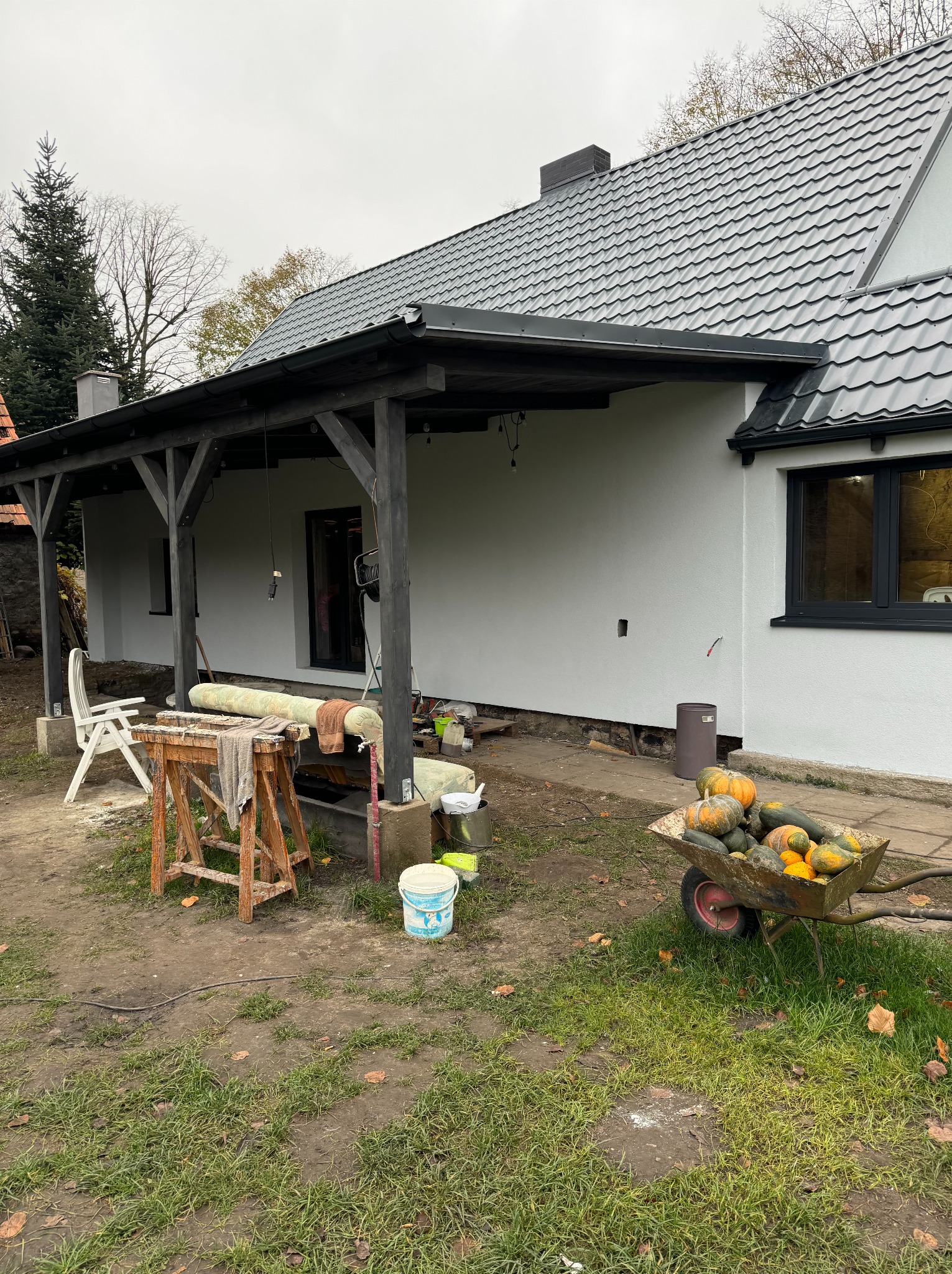 Biała elewacja domu z szarą dachówką i drewnianą pergolą. Wózek z dyniami, narzędzia i materiały budowlane sugerują prace wykończeniowe.