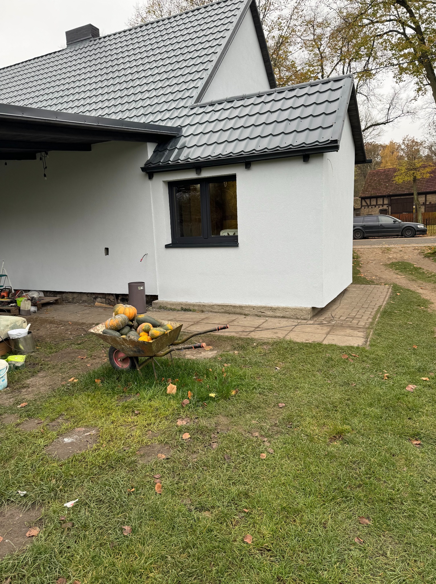 Biała elewacja domu z ciemną stolarką okienną i dachem. W tle wózek z dyniami, trawnik i budynek gospodarczy. Jesienna sceneria.