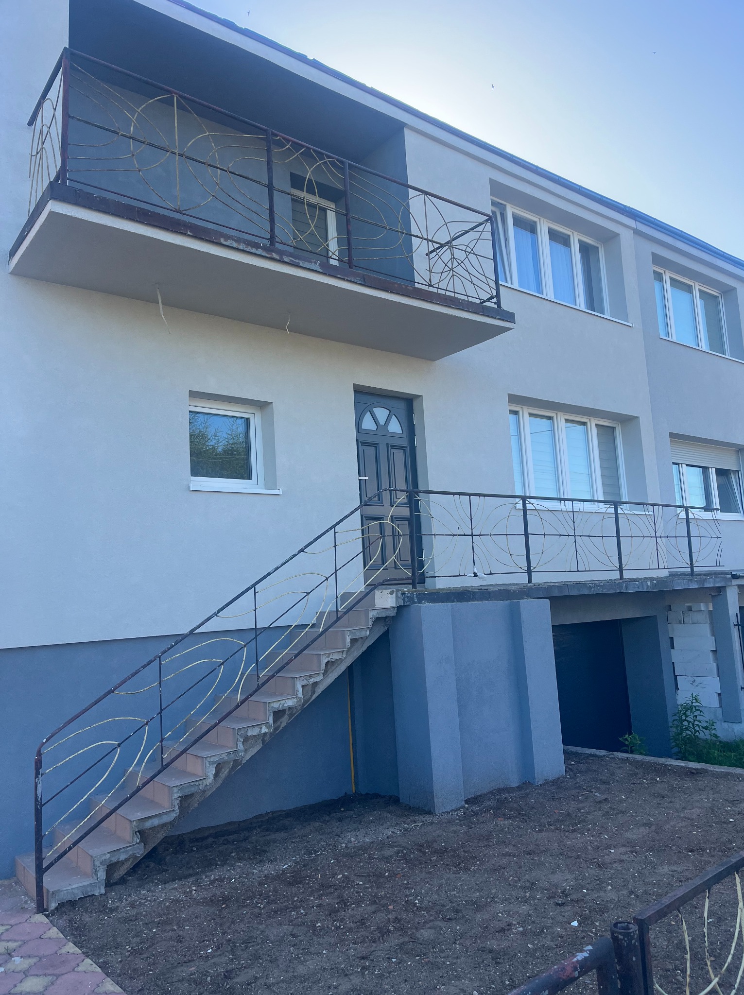 Dwukondygnacyjny dom z balkonem i zewnętrznymi schodami z ozdobnymi, metalowymi balustradami. Elewacja w odcieniach szarości, okna z białymi ramami. Ujęcie z dołu.