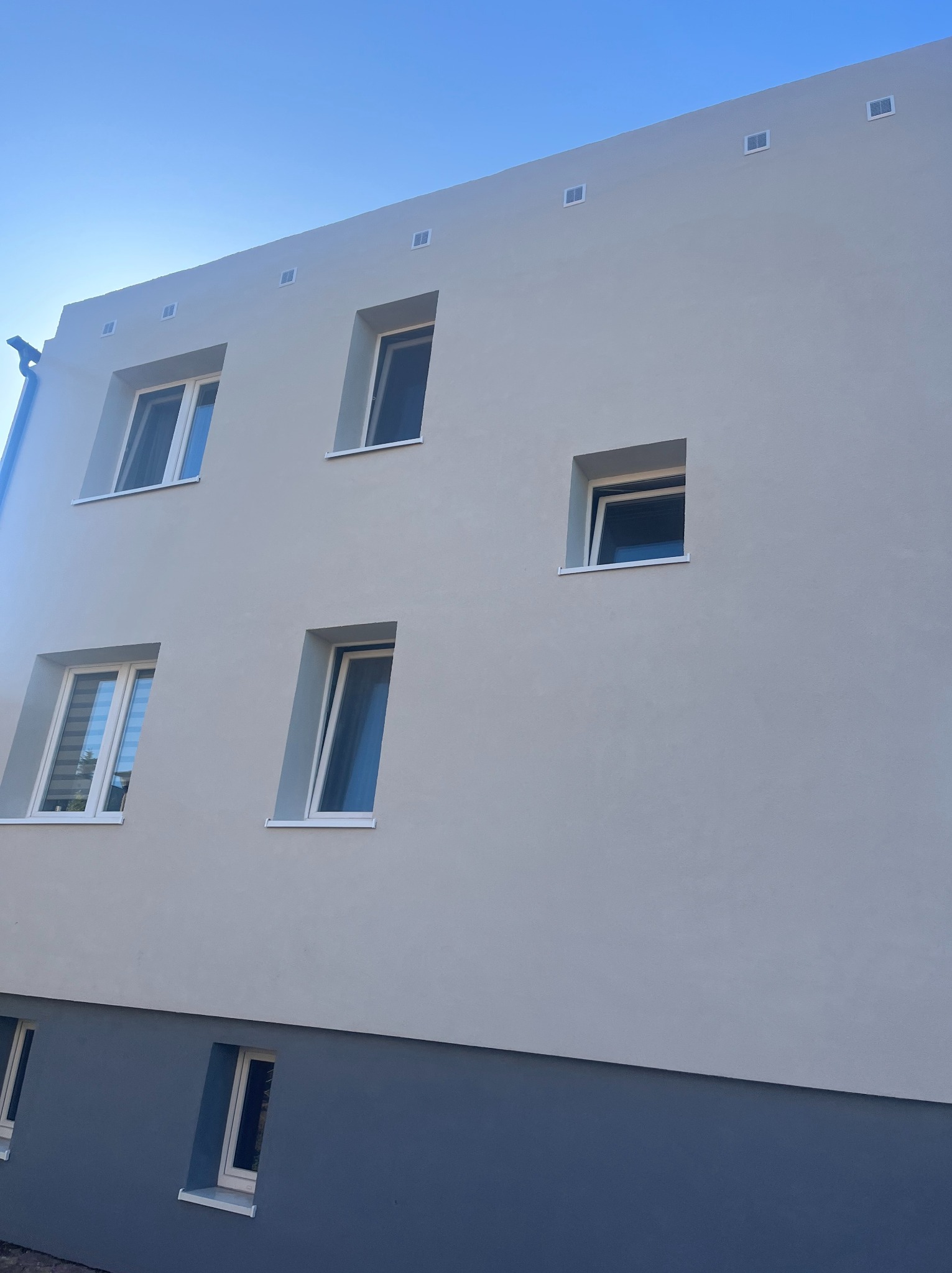 Elewacja budynku po tynkowaniu, z oknami w białych ramach i widocznymi kratkami wentylacyjnymi pod dachem, na tle błękitnego nieba.