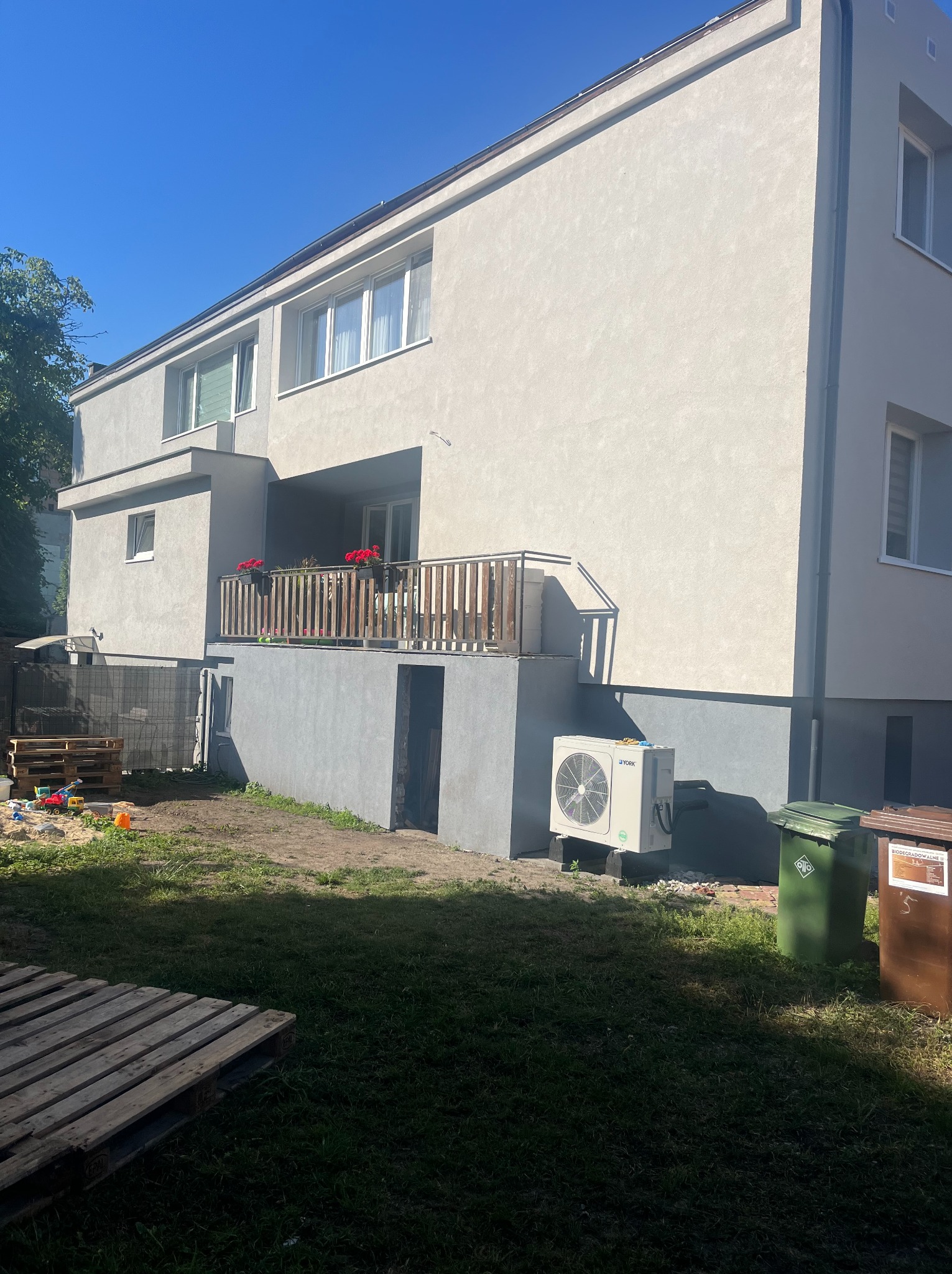 Dwupiętrowy dom z szarą elewacją, balkonem z drewnianą balustradą i klimatyzatorem York na zewnątrz. Widoczne kosze na śmieci i plac zabaw.