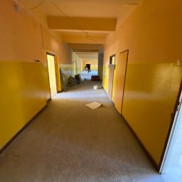 MjHorizonLine Sp Z o.o. - Korytarz w trakcie odświeżania: ściany pomalowane w dolnej części na żółto, w górnej na pomarańczowo, widoczne ślady prac malarskich na podłodze, w tle ustawione drabiny malarskie i wiadro z farbą.