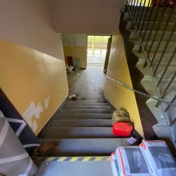 MjHorizonLine Sp Z o.o. - Klatka schodowa w trakcie remontu: żółta ściana z niedomalowanymi fragmentami, worki zaprawy tynkarskiej na schodach, kanistry i resztki materiałów budowlanych. Widoczne zabezpieczenia...