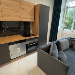 MjHorizonLine Sp Z o.o. - Nowoczesna kuchnia z drewnianymi frontami szafek, czarnym blatem i marmurową ścianą, widok na szarą zabudowę i narożnik z poduszkami, jasna marmurowa podłoga, okno z zielonymi zasłonami w tle.