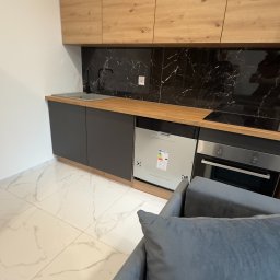MjHorizonLine Sp Z o.o. - Nowoczesna kuchnia z drewnianymi szafkami górnymi, szarymi szafkami dolnymi i czarnym marmurowym backsplashiem, widok z perspektywy sofy.