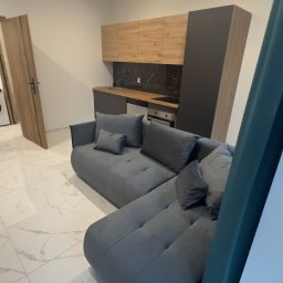 MjHorizonLine Sp Z o.o. - Nowoczesna kuchnia z szarymi szafkami i drewnianymi frontami w Bytomiu, widoczna z perspektywy narożnej sofy w odcieniach szarości na białej marmurowej podłodze.